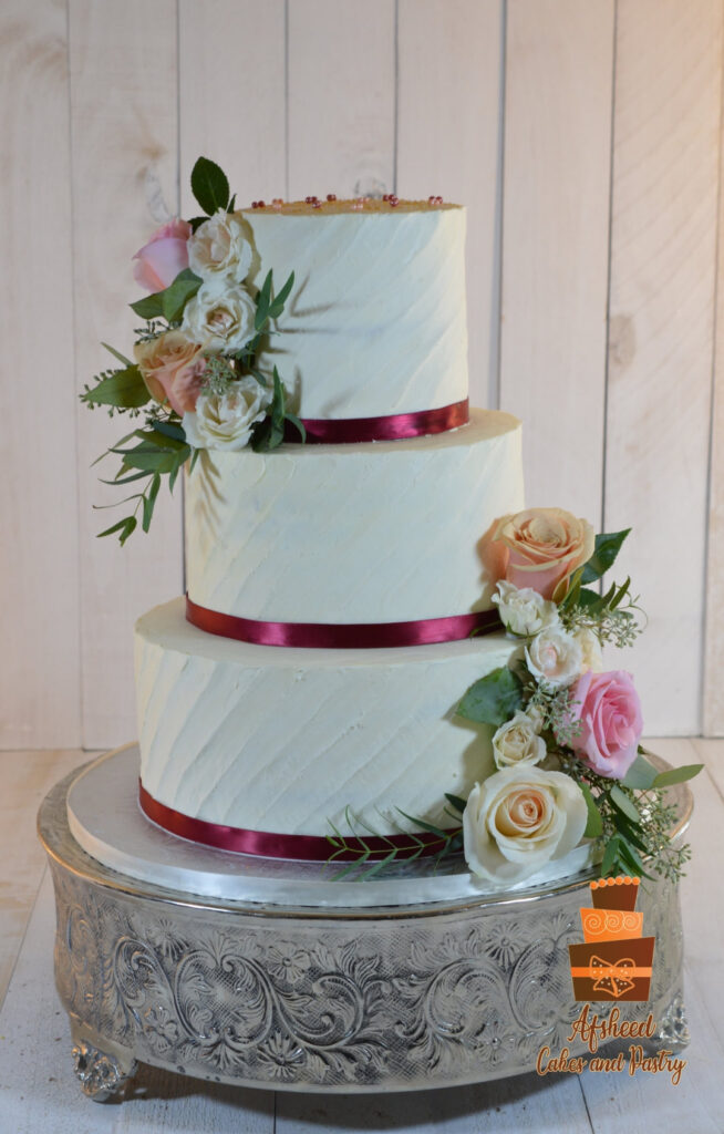 Afsheed Cakes custom wedding cake in Calgary. Elegant three-tier textured ivory buttercream cake with burgundy ribbon and fresh floral cascade of pink and peach roses.