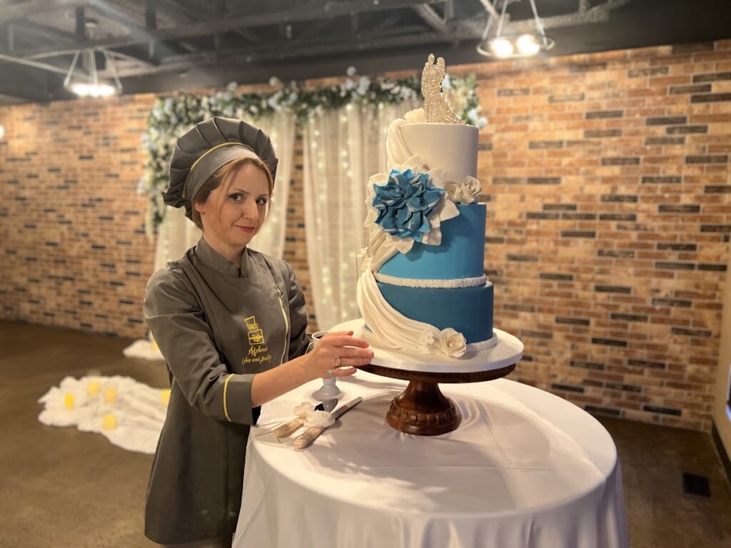 A three-tier blue and white wedding cake with elegant draped fondant and floral decorations, displayed on a wooden cake stand by a pastry chef from Afsheed Cakes and Pastry in Calgary.