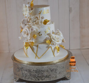 Luxury gold geometric wedding cakes Calgary - Three-tier white fondant cake featuring gold geometric line patterns, gold leaf accents, and a lush cascade of white and cream sugar roses with gold-painted leaves by Afsheed cakes, in Calgary for couples seeking a modern, artistic, and glamorous custom wedding cake.