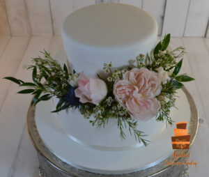 Custom outdoor wedding cakes Calgary - Two-tier smooth white fondant cake topped with soft pink peonies, blue thistles, and delicate greenery delivered to Bow Valley Ranch Restaurant in Fish Creek Provincial Park by Afsheed cakes, in Calgary for couples planning a garden-style or rustic luxury wedding.