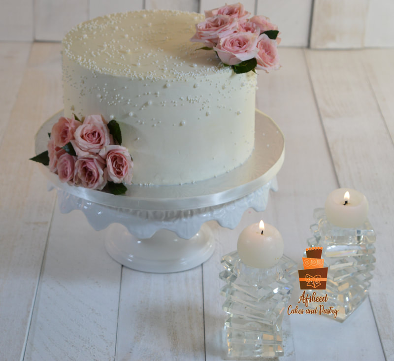 Custom pearl wedding cakes Calgary - Single-tier ivory buttercream cake delicately decorated with white edible pearls and two clusters of fresh pink roses, displayed next to elegant crystal candle holders by Afsheed cakes, in Calgary for couples seeking a romantic, intimate, and highest rated custom bakery experience.