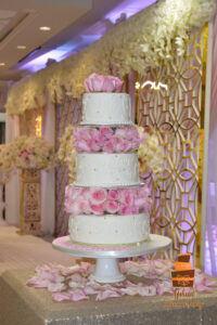 Extravagant multi-tier wedding cakes Calgary - Tall five-tier white fondant cake featuring intricate royal icing scrollwork, gold accents, and lush separators made of fresh pink roses by Afsheed cakes, in Calgary for couples planning a grand, luxurious, and high-end wedding celebration.