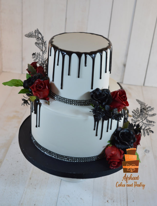 Gothic Wedding cake Calgary - Bold two-tier white fondant cake featuring a dark chocolate drip, black rhinestone bands, and a striking arrangement of deep red and black sugar roses with dark fern accents by Afsheed cakes, in Calgary for couples seeking a dramatic, gothic-glam, or modern luxury wedding cake from the highest rated custom bakery.