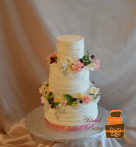 Rustic buttercream wedding cakes Calgary - Three-tier white cake with rough-textured horizontal buttercream ridges, encircled by rings of delicate pink and white blossoms and dark berries, which traveled from Calgary to Whitefish Montana by Afsheed cakes, in Calgary for couples planning a destination, rustic-chic, or mountain-inspired luxury wedding.