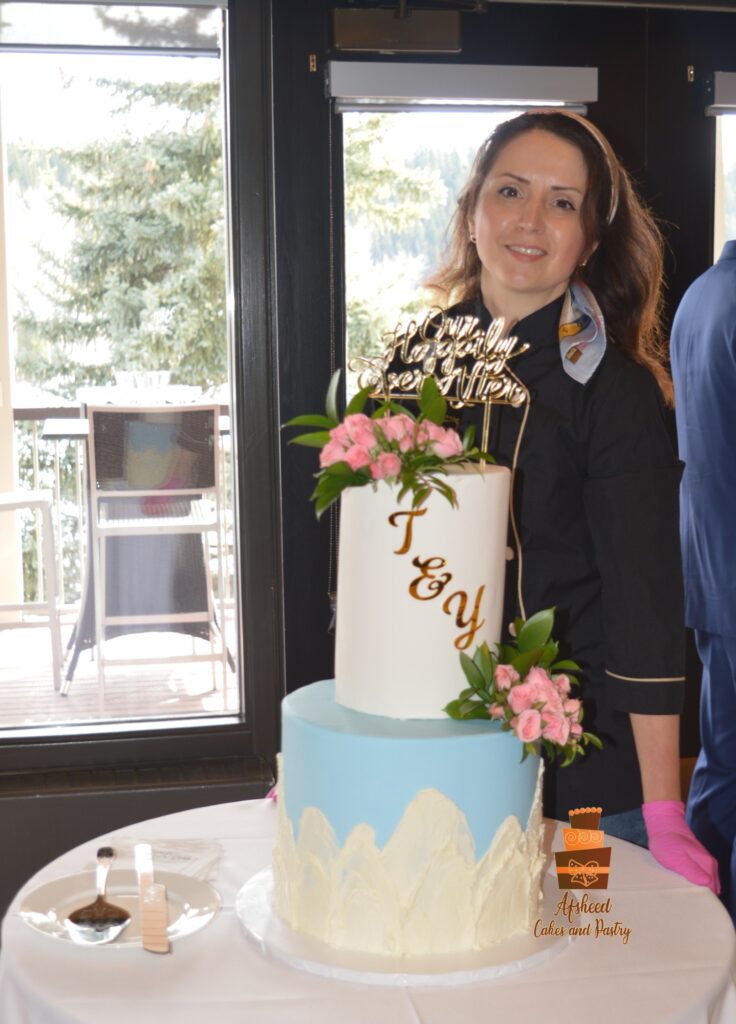 Tall two-tier buttercream wedding cake with fresh blush pink roses and leaves, decorated with custom cake topper, filled with chocolate ganache and decorated with mountain patterns on the side, delivered and setup to Calgary waterfront park, by Afsheed Cakes